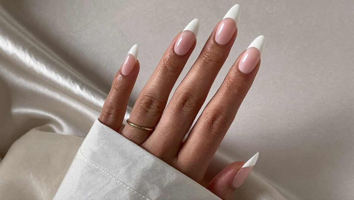 Almond nails with a sheer pink base and crisp white French tips emerging from a white shirt cuff against grey satin