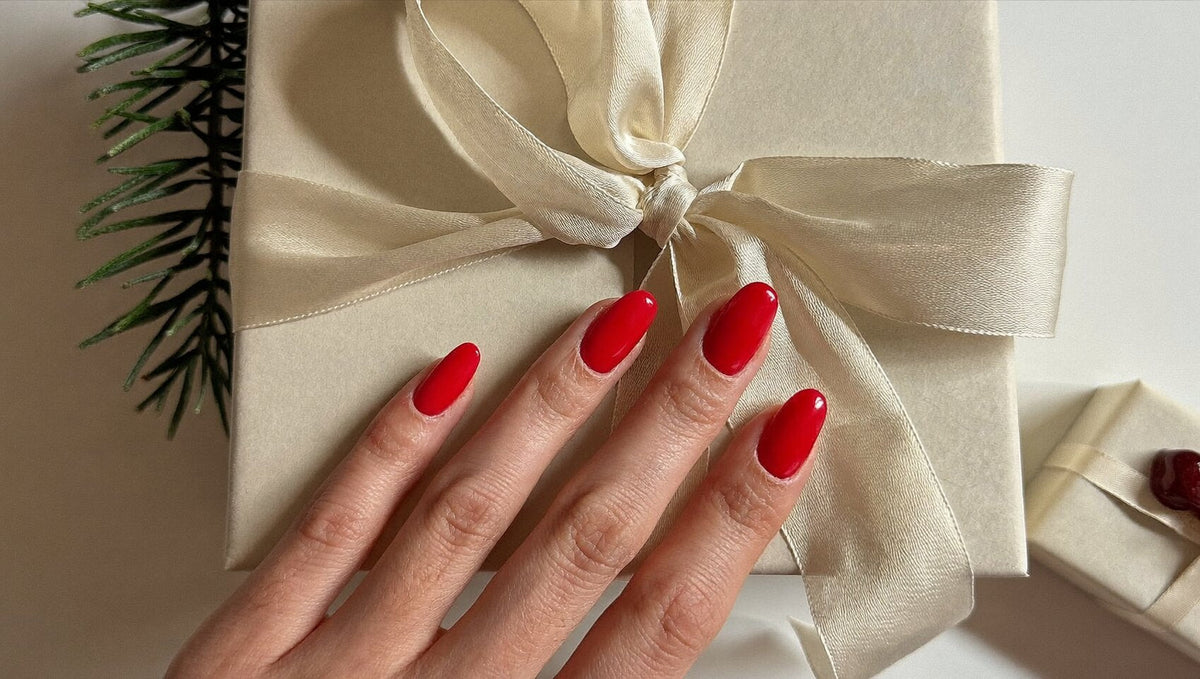 A hand with glossy red almond nails resting on a gift box wrapped in ivory paper with a champagne silk ribbon and a sprig of pine