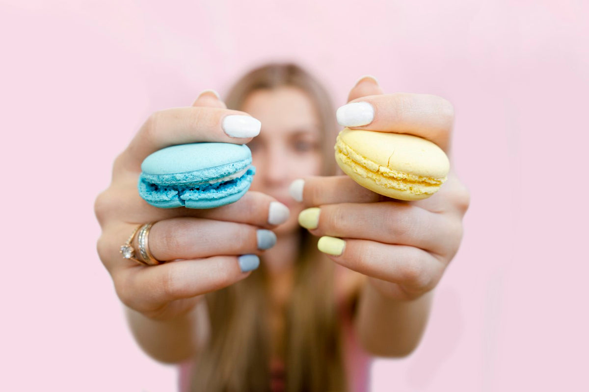Hands with pastel blue, white and yellow nails holding two macarons against a pink background