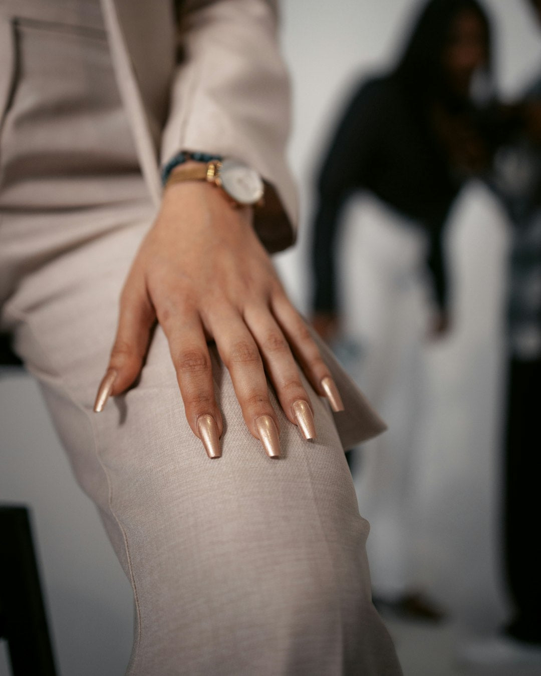 Hand with long gold chrome almond nails resting on a beige trouser suit