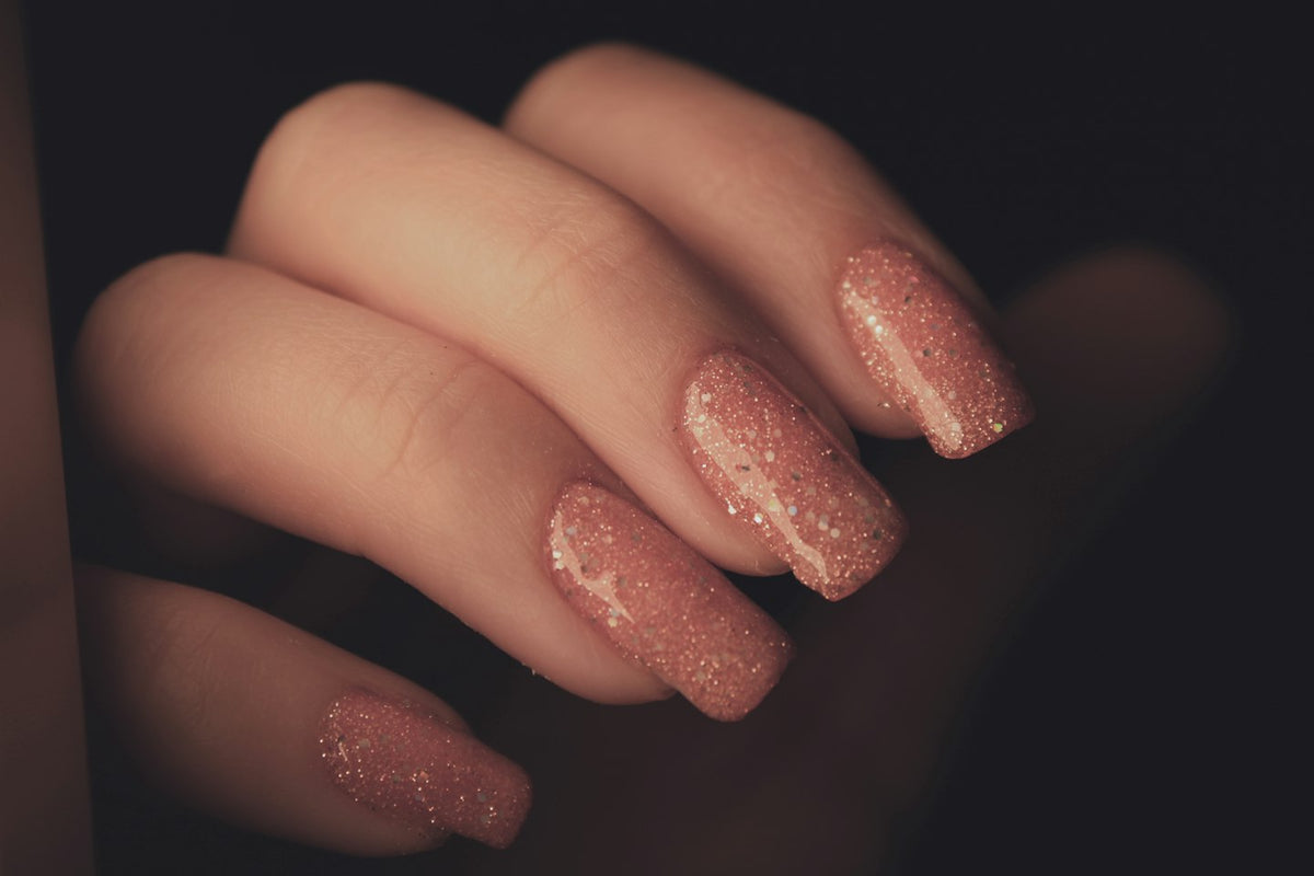 Hand with long square nails in rose gold glitter gel against a dark background
