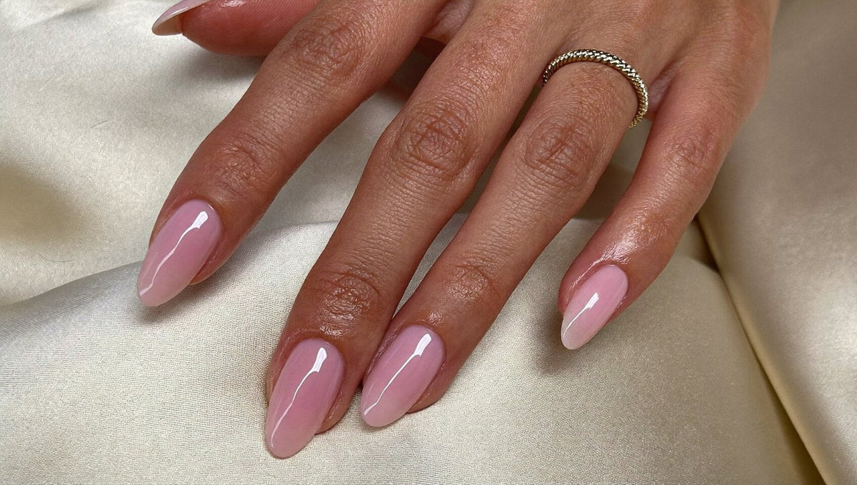 Hand with short almond nails in a glossy milky pink and a thin gold ring resting on cream silk