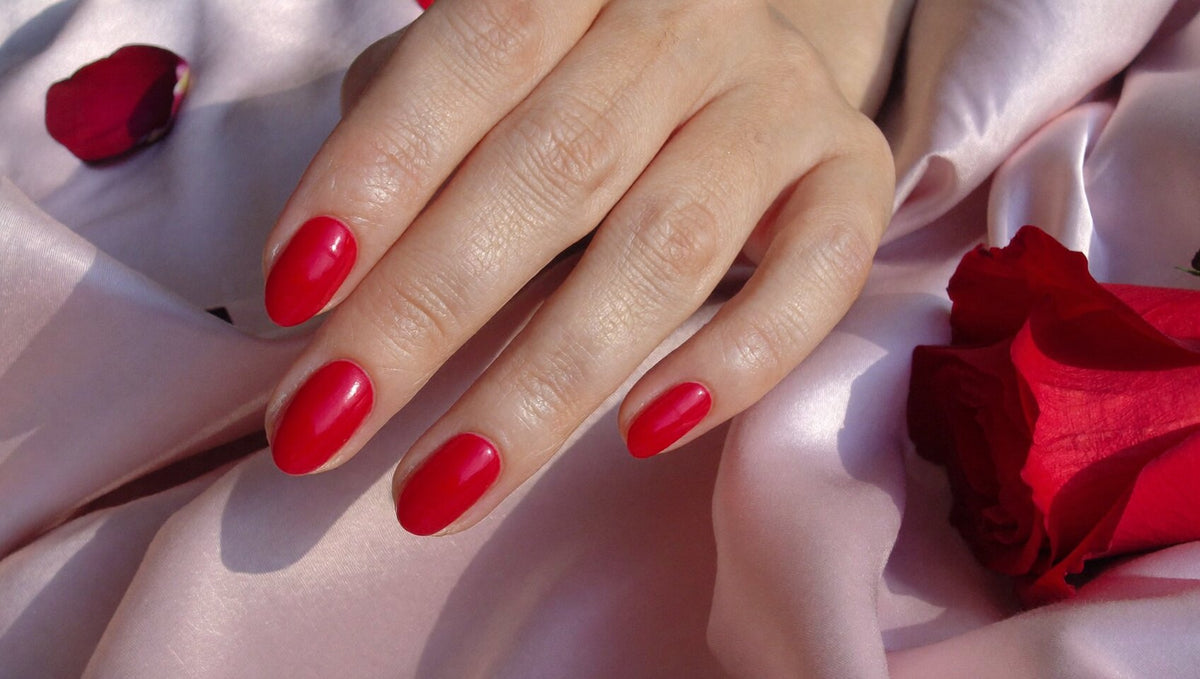 Hand with short almond nails in a glossy cherry red resting on pale pink silk beside a red rose