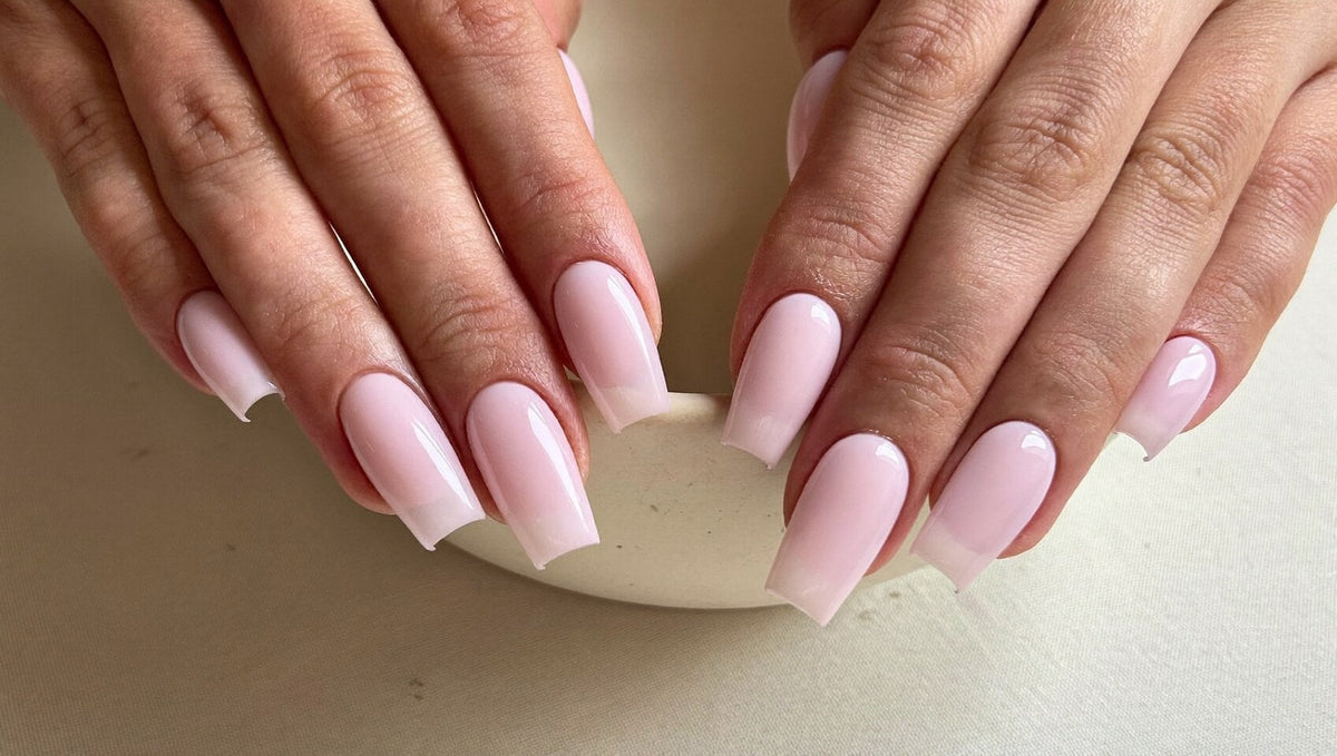 Two hands with long square nails in a smooth, glossy milky pink builder gel resting on the rim of a cream ceramic bowl
