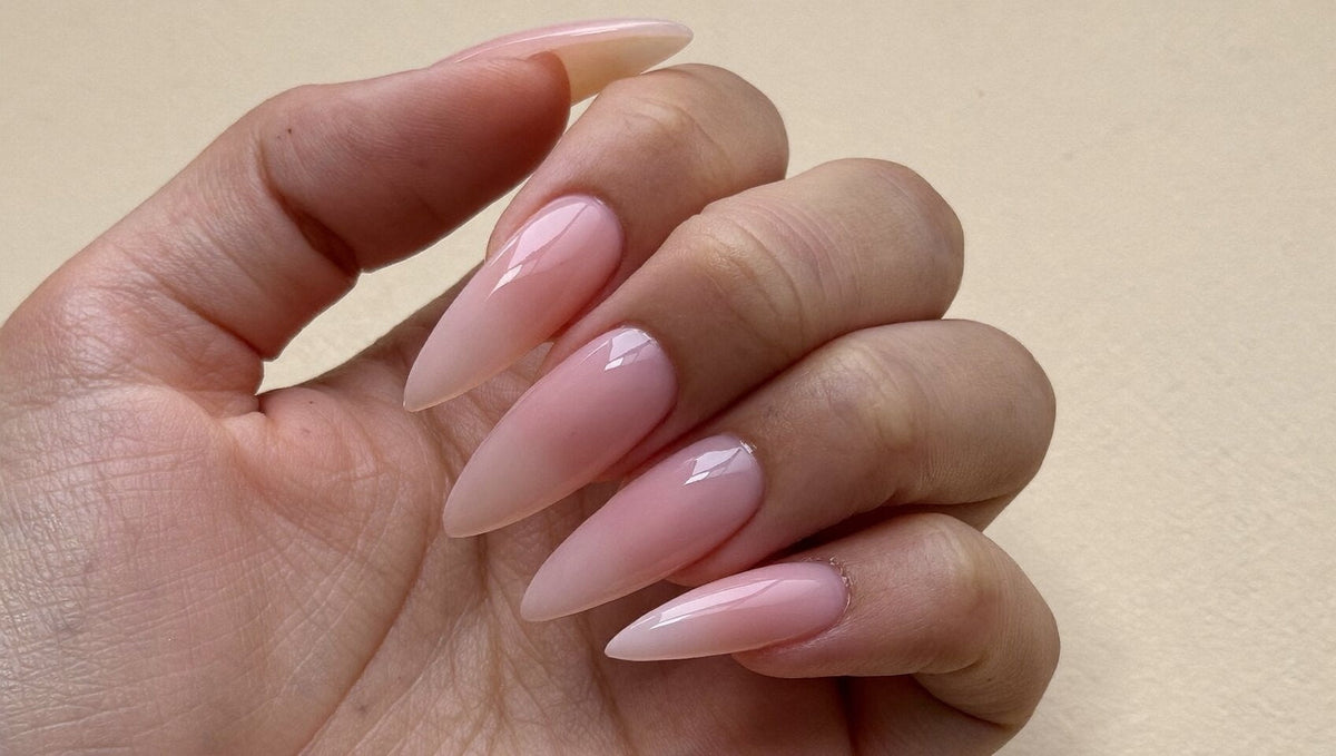 Hand with long almond nail extensions in a smooth, glossy milky pink builder gel on a warm cream background