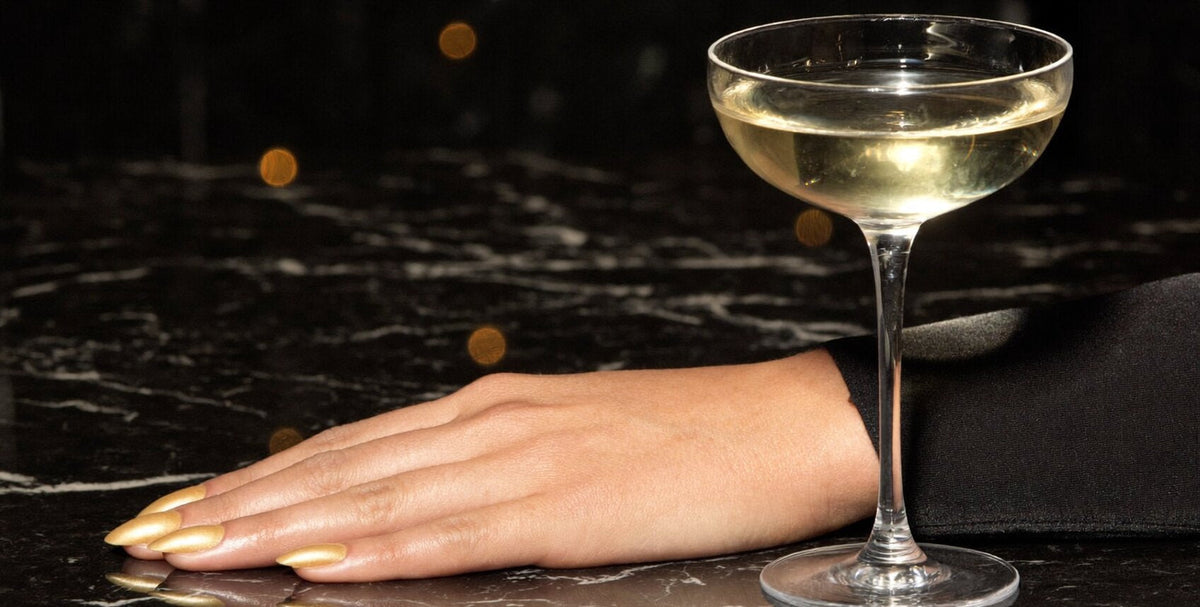 Hand with glossy liquid gold almond nails resting on black marble beside a champagne coupe, warm golden lights in the background
