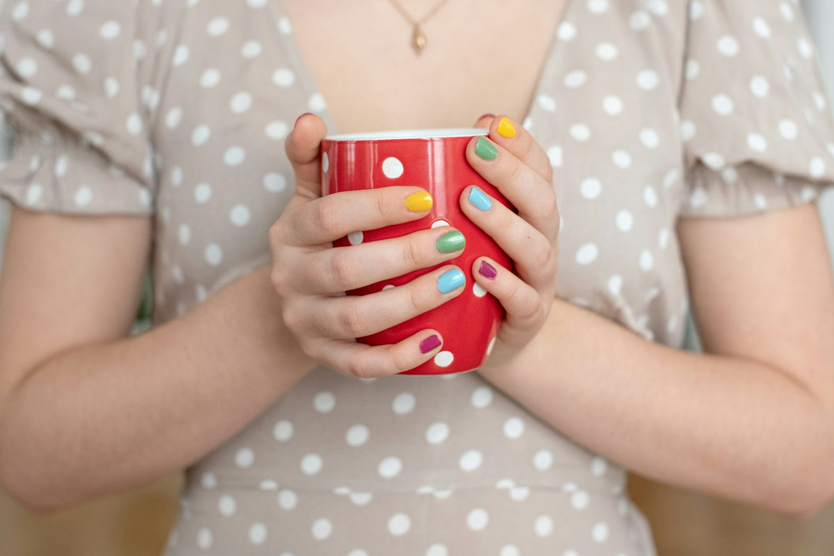 Hands with multicoloured nails holding a red polka dot mug against a polka dot dress
