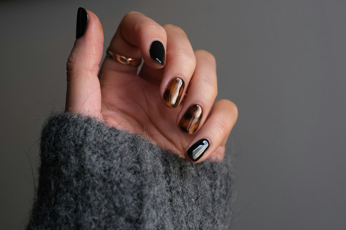 Hand with short glossy black and tortoiseshell nails and a gold ring against a grey knit sleeve