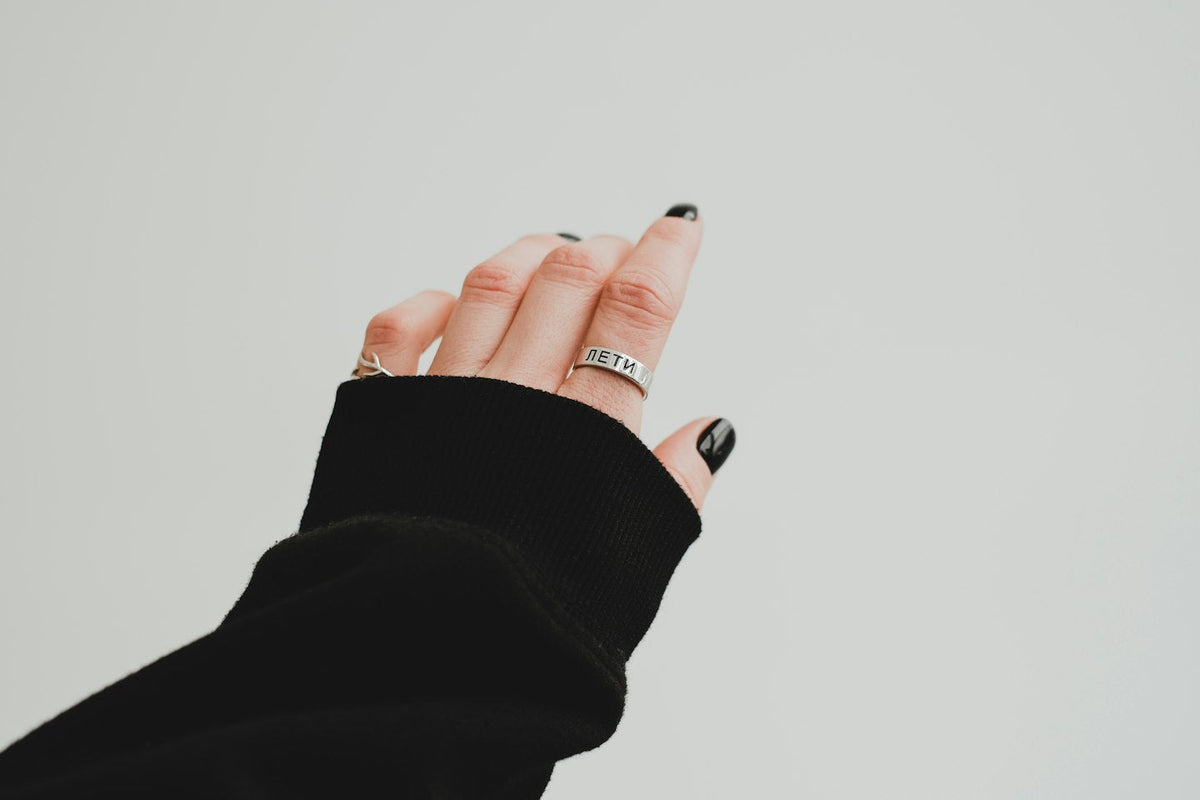 Hand with short glossy black nails and a silver ring, black sleeve, pale grey background