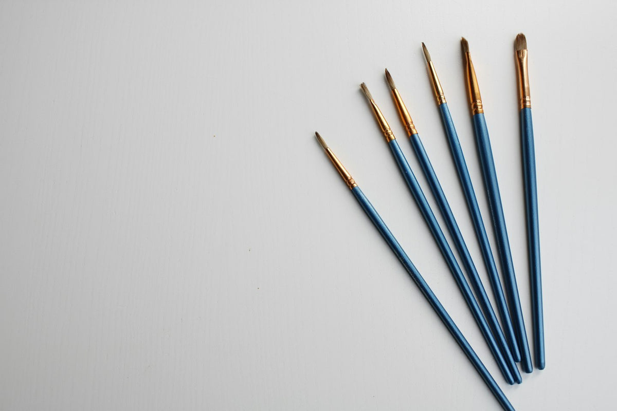 Six fine-tipped brushes laid side by side on a white surface
