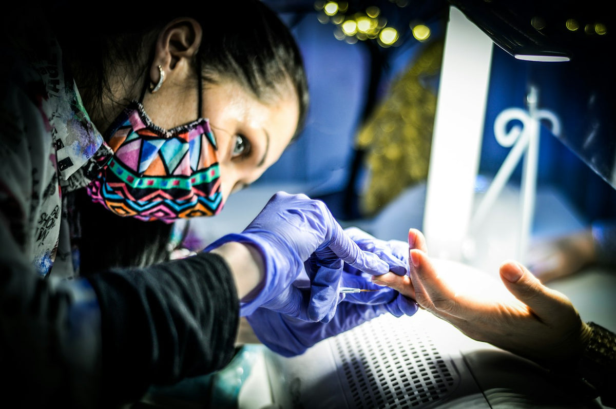 Nail technician in a face mask and purple gloves working on a client's nails under a lamp