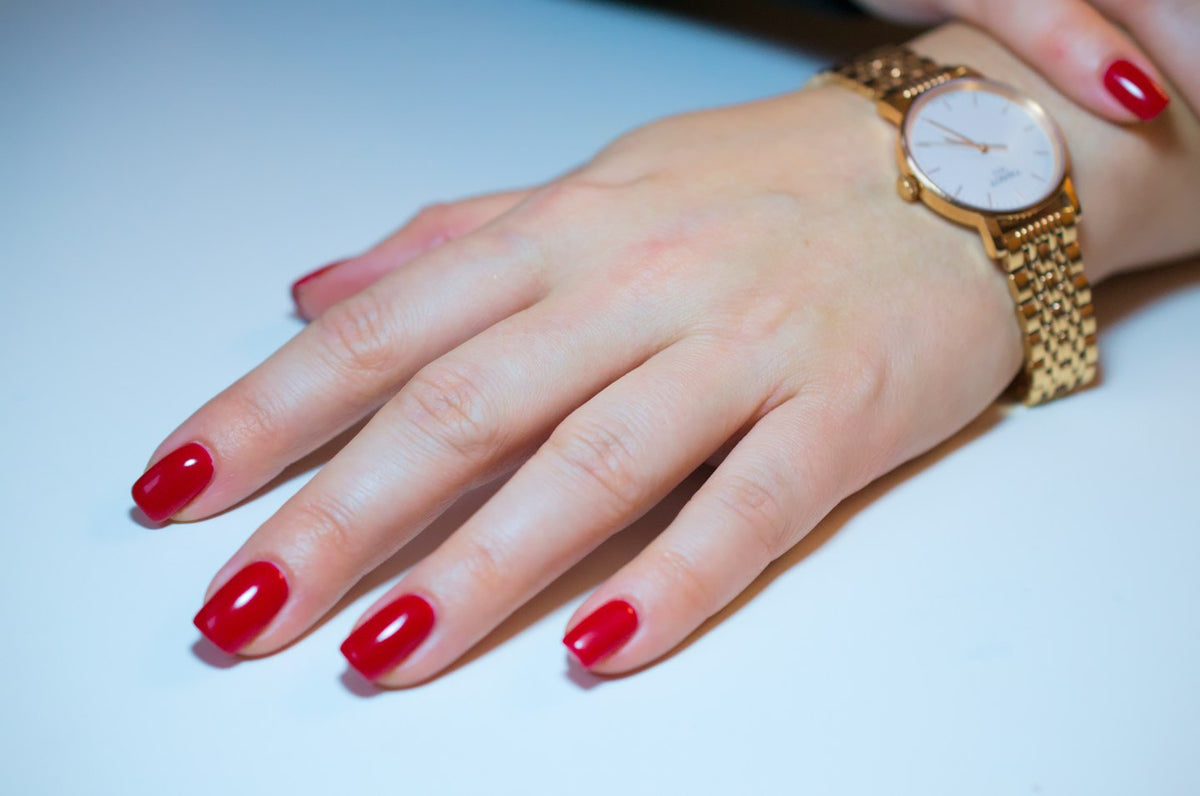 Hand with short glossy red nails and a gold watch on a white surface