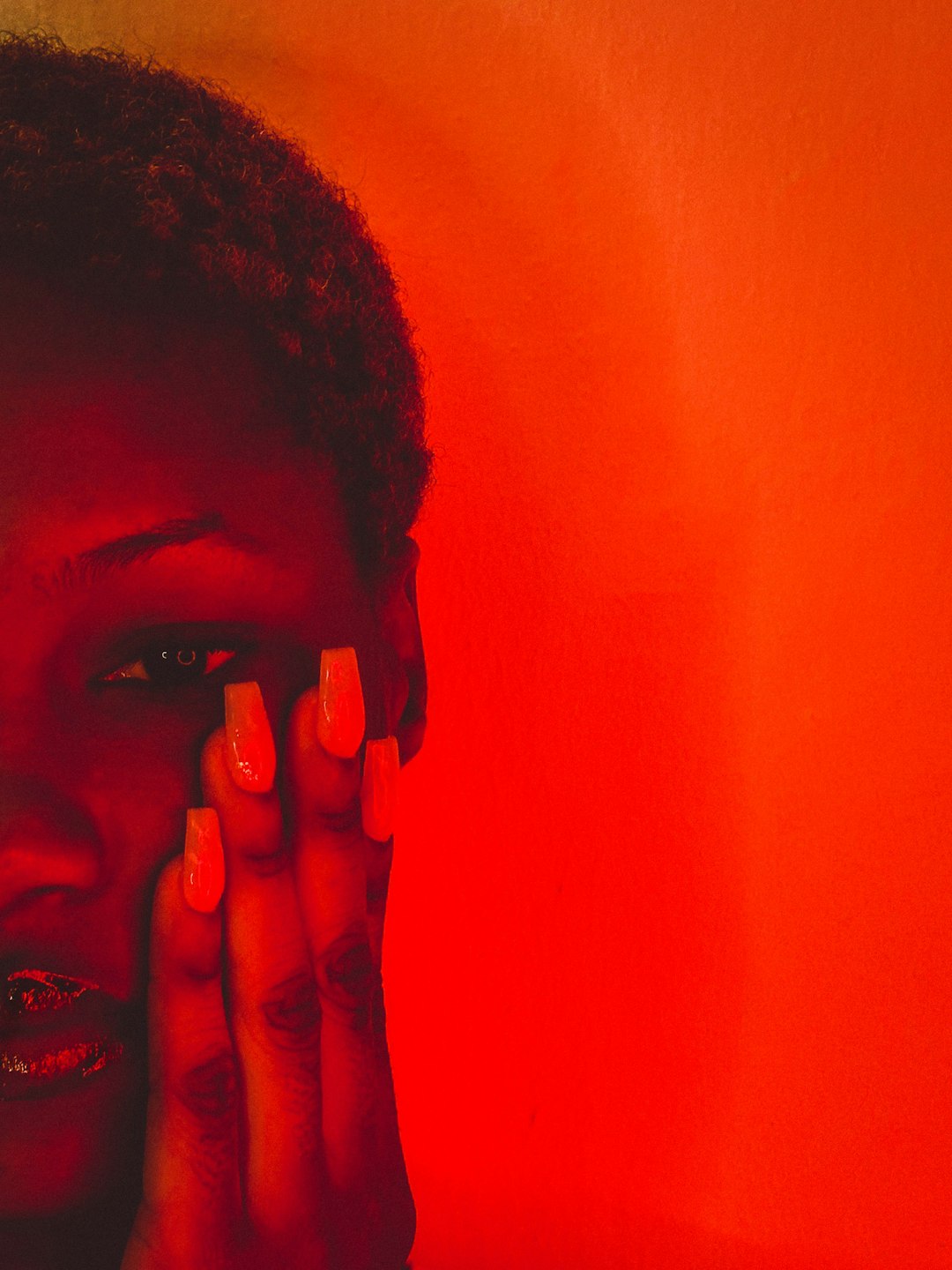 Hand with long bright orange nails held to the face against an orange-red background