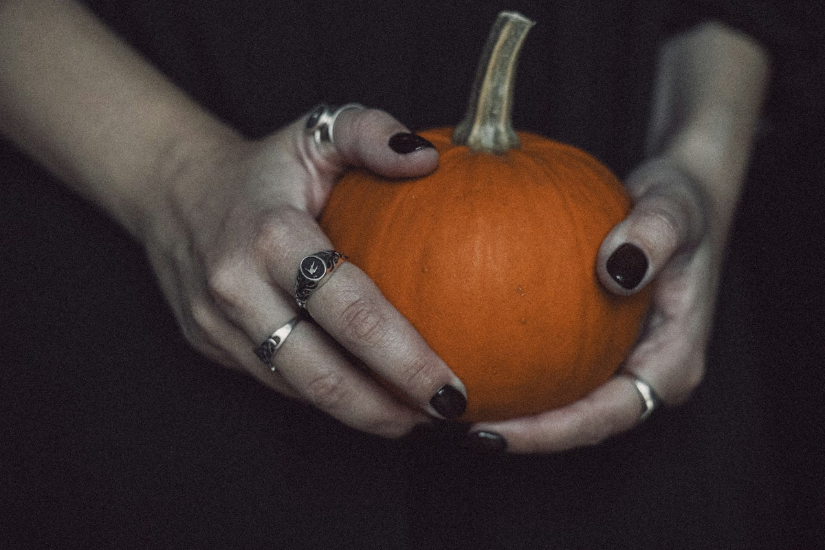 Mains aux ongles prune foncé et bagues argentées tenant une petite citrouille orange sur fond sombre