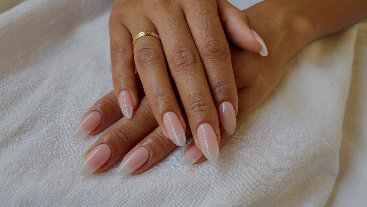 Two hands resting on ivory linen, short natural almond nails strengthened with a sheer milky pink gel, one thin gold ring